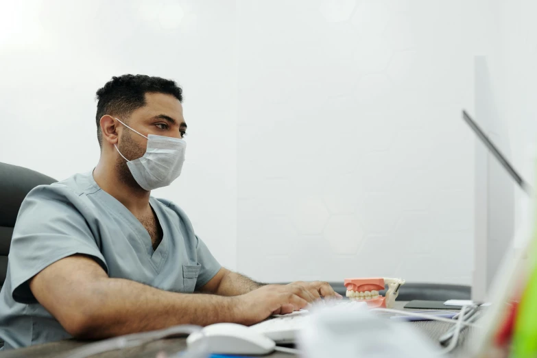 a male doctor in scrubs sits at a desk wearing a medical mask