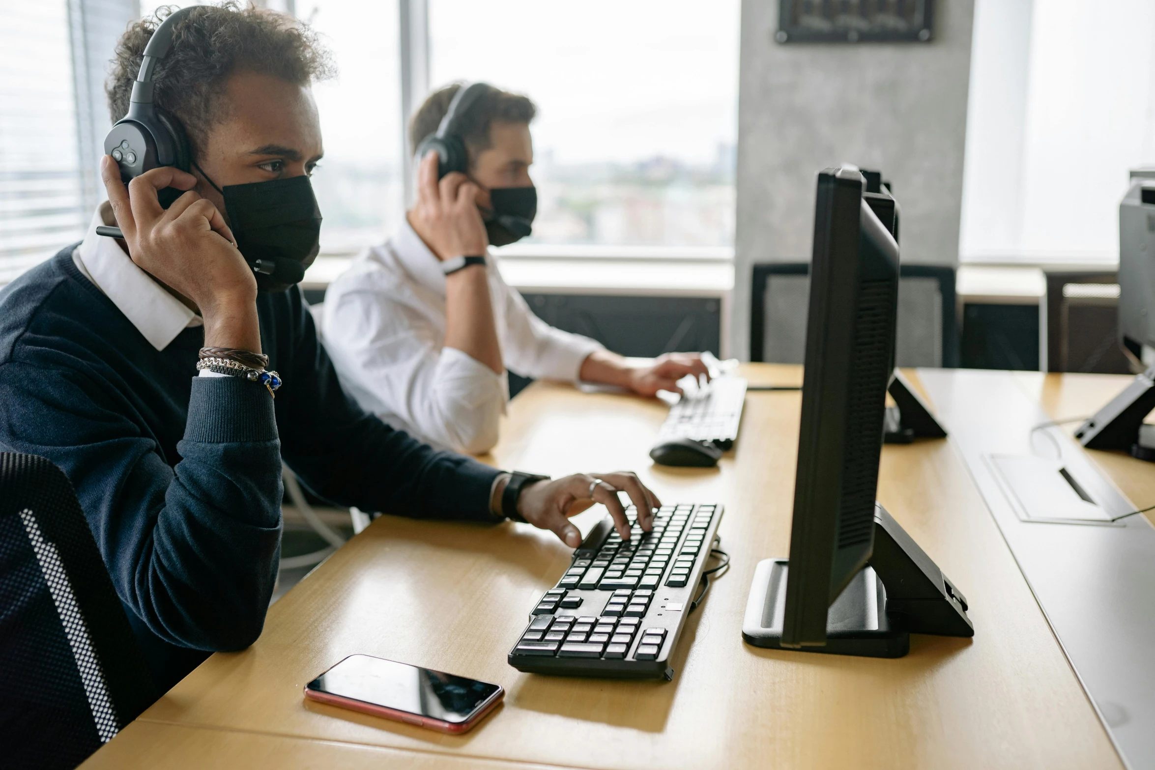 a man wearing a mask sitting at a computer while on the phone