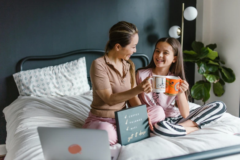 a woman and a girl sit on a bed and look at an open laptop