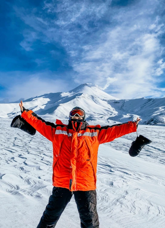 a man in an orange jacket and black pants stands on a snow covered hill with his arms out