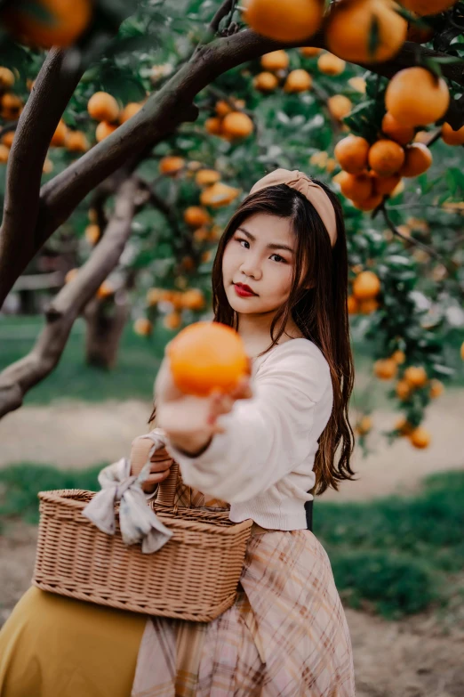 girl in white shirt holding orange and standing next to tree