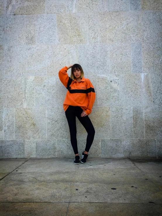 a woman wearing a long orange shirt and black pants leaning against a wall