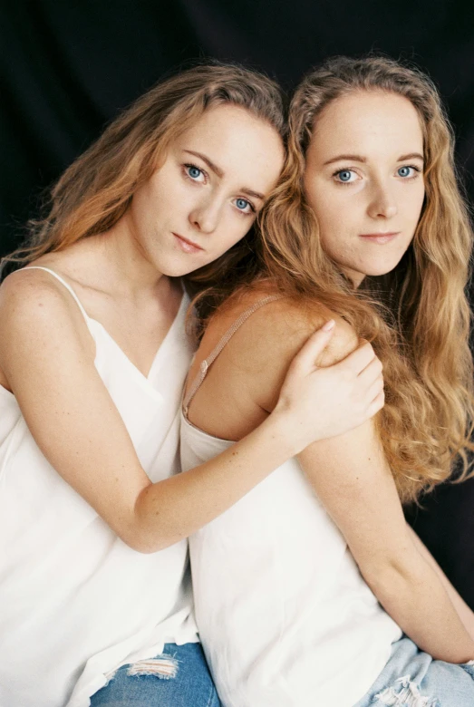 two girls hugging on black background one wearing a white tank top and the other wearing blue denim jeans