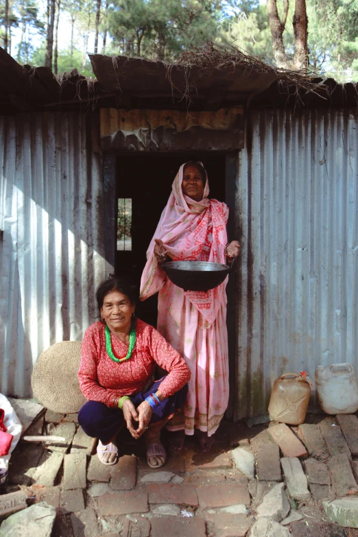 two women sitting down outside a building cooking food