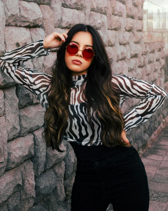 a woman with long hair wearing sunglasses standing by a stone wall