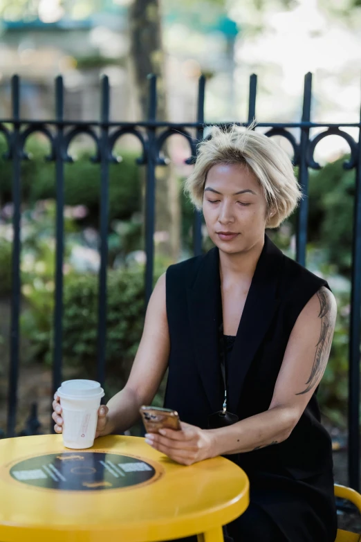 a woman sitting at a table holding a cellphone and looking at a cup of coffee