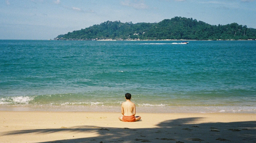 a man is sitting on the beach with a boat out to sea