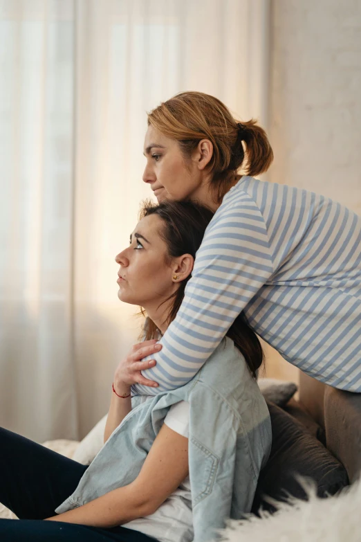 two women sit on a couch, each holding their hand over their shoulder