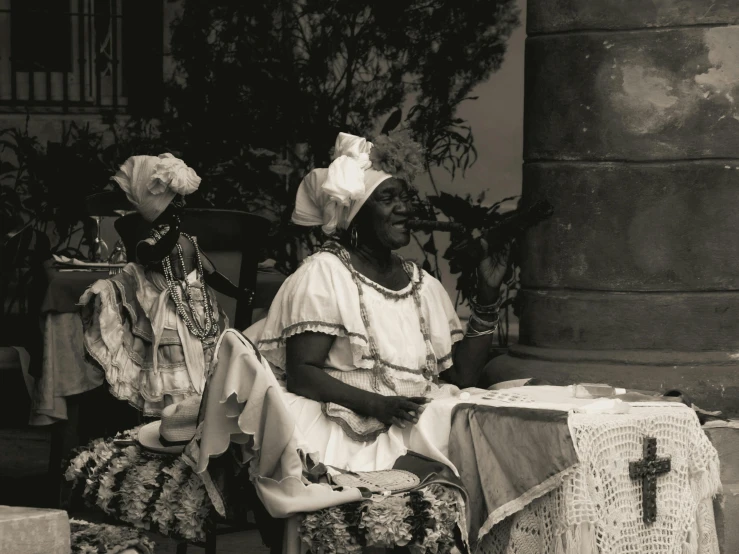 two people sit at an outdoor table wearing dresses and accessories