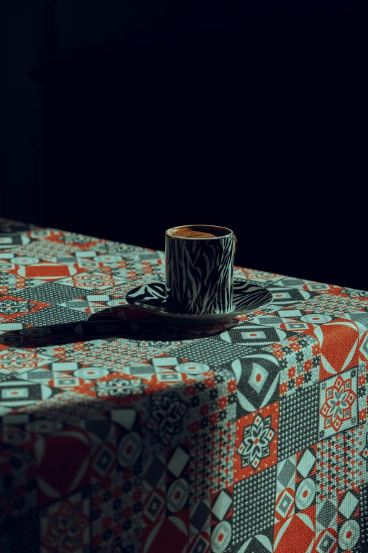 an orange, grey and white table cloth has a black, silver and red patterned cup on top