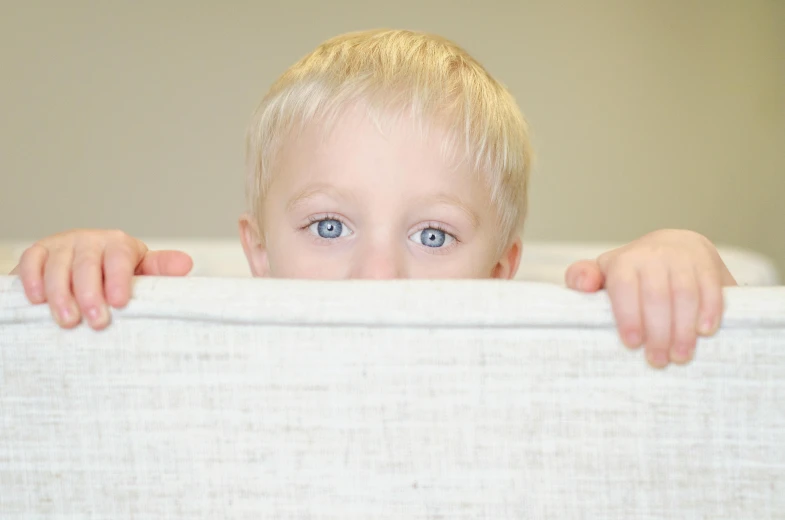 a little boy standing behind a white bed with his arms on the head and eyes wide open