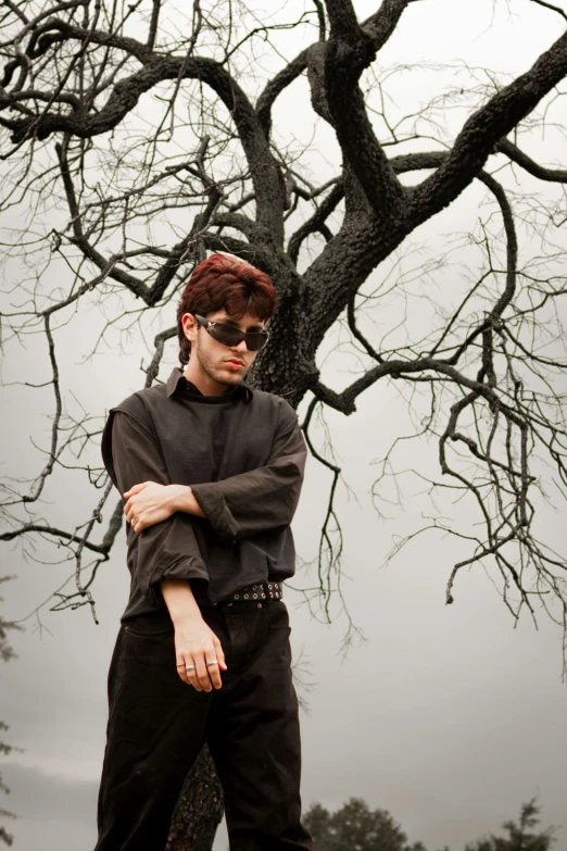 man wearing shades, shirt and pants standing next to large tree