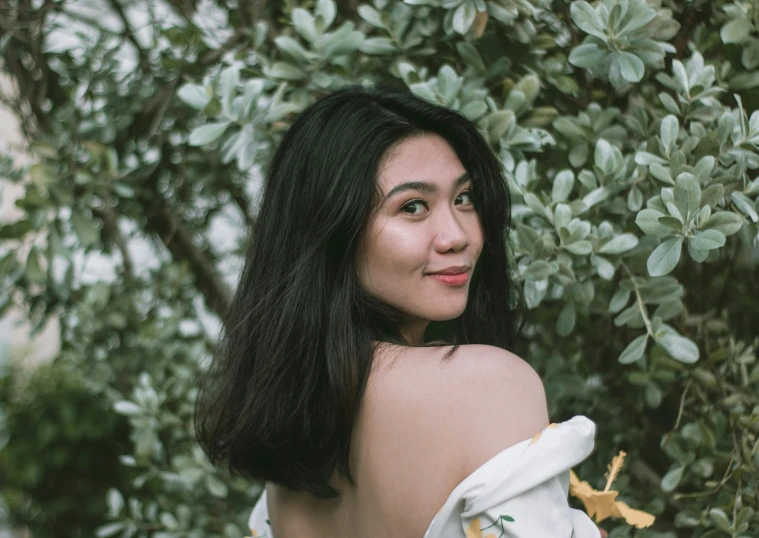 a young woman with dark hair, wearing white off the shoulder dress and looking over her shoulder