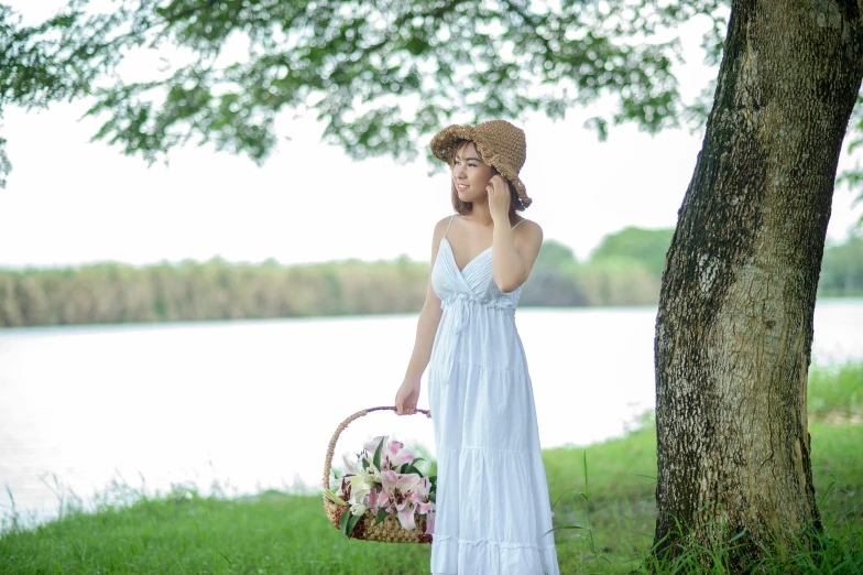 a woman in a dress and straw hat next to a tree holding a basket