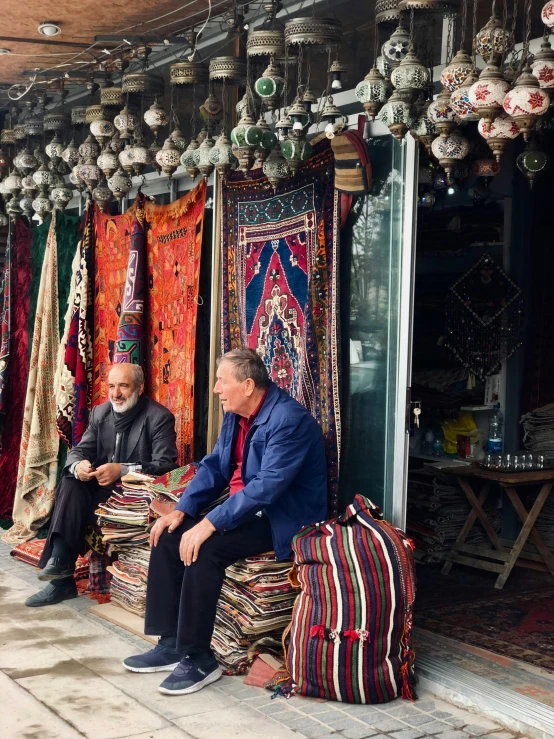 two men sitting in front of a shop with colorful carpets
