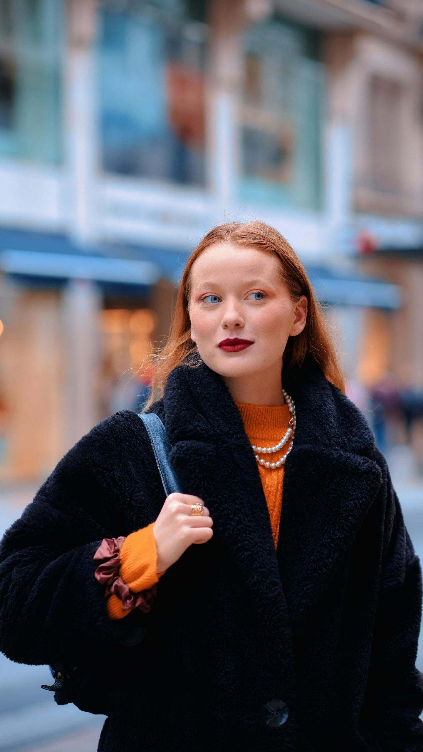a red headed woman in an orange sweater, black coat and a pearl necklace, on a city street