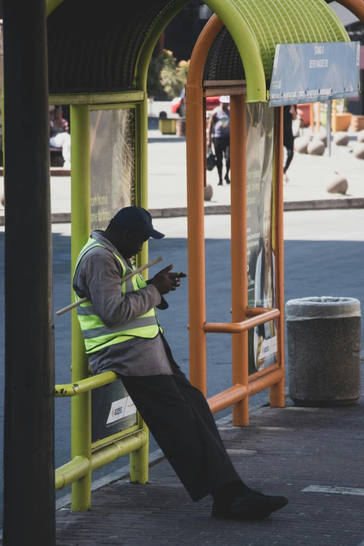 a man in an orange vest sitting on a bench in a bus station while looking at his cell phone