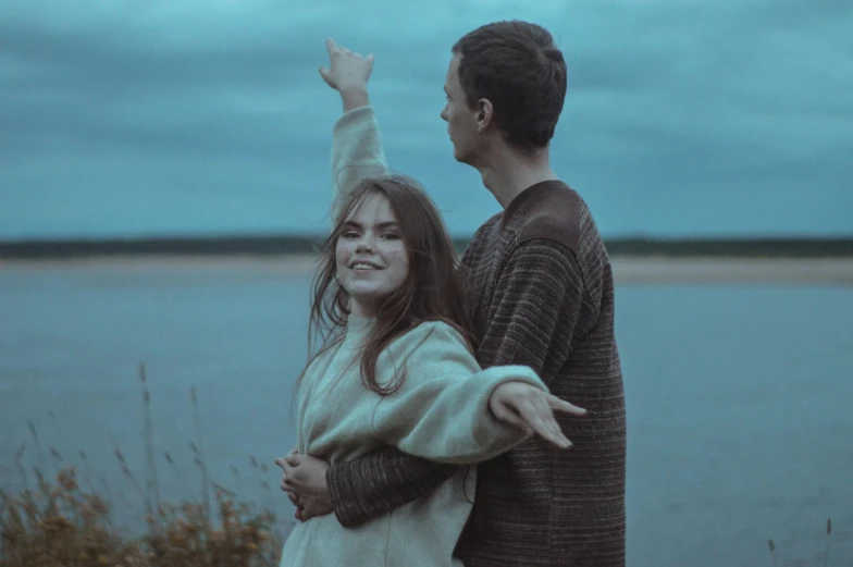 two people, one with her arms outstretched and one holding their head down, are standing by the water at twilight