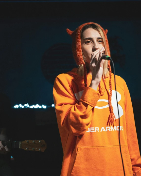 the woman is singing on stage in an orange sweatshirt