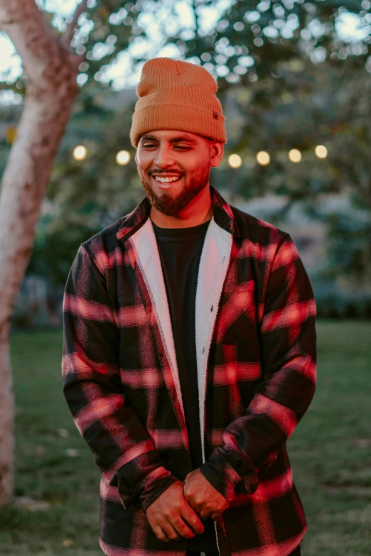a bearded man standing in the grass, wearing a plaid jacket
