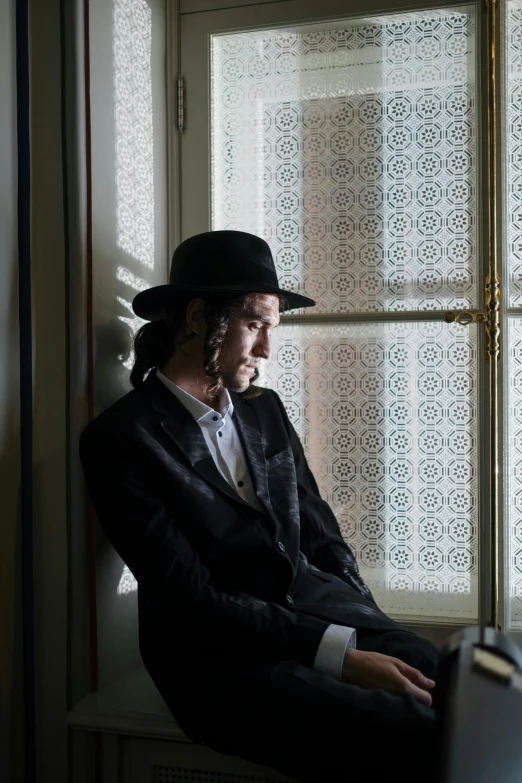 a man wearing a black hat and suit sits by a window