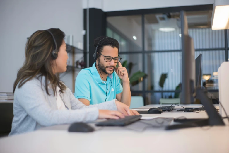 two people sitting at a computer talking on their cell phones