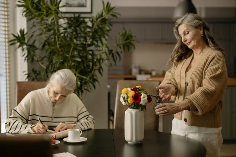 a woman prepares flowers in a vase with another woman seated at the table