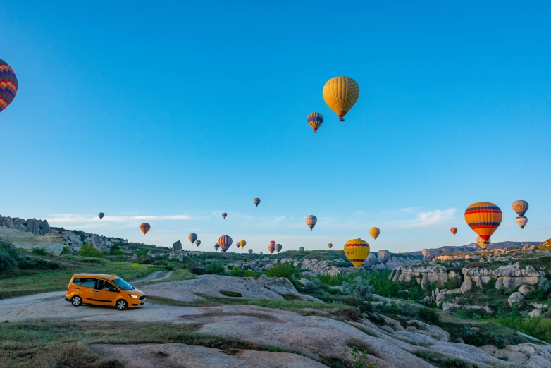 several  air balloons are flying above a yellow car on the side of a dirt road