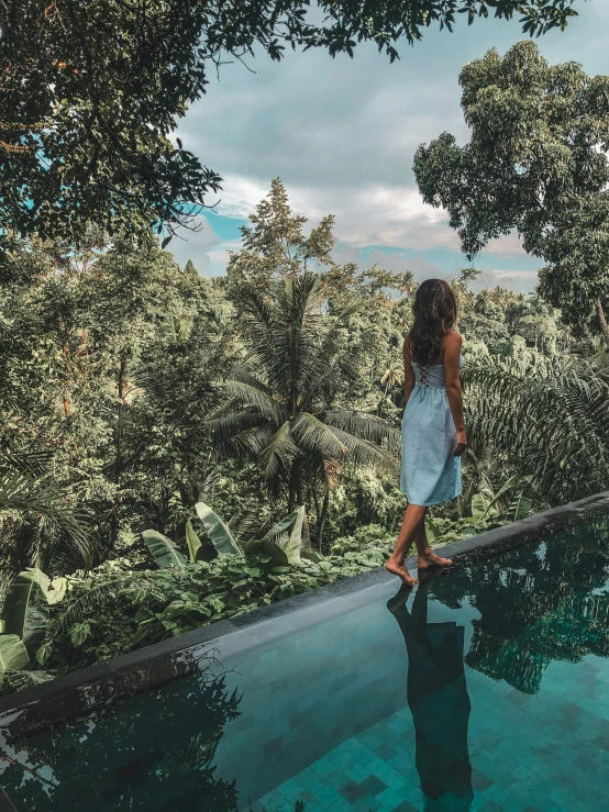 a woman in blue and white dress standing by a pool with trees