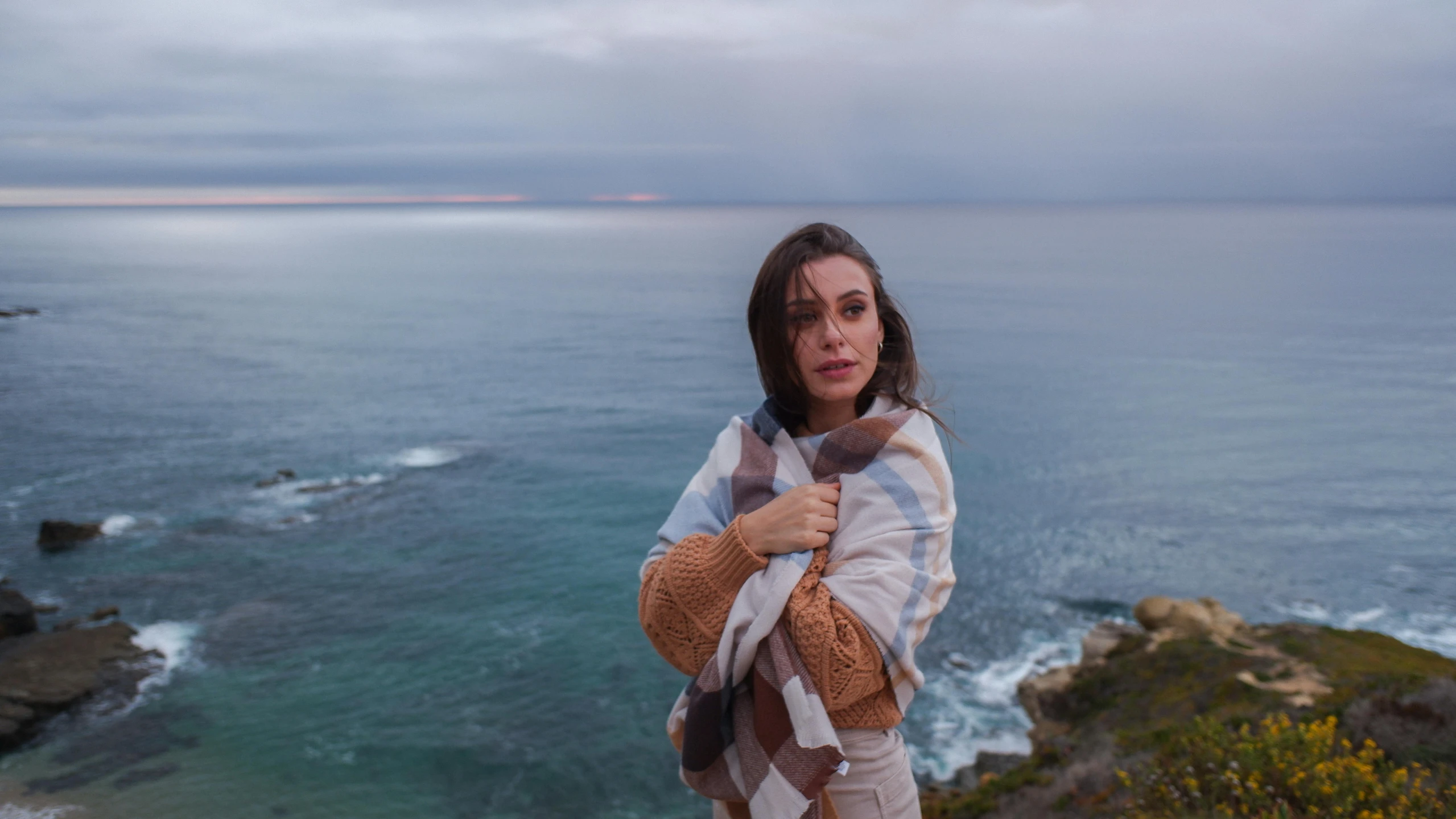 a woman poses near an ocean, while wearing a scarf