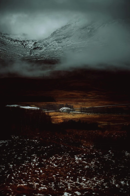 the clouds gather over a small creek that's surrounded by a snow covered mountain