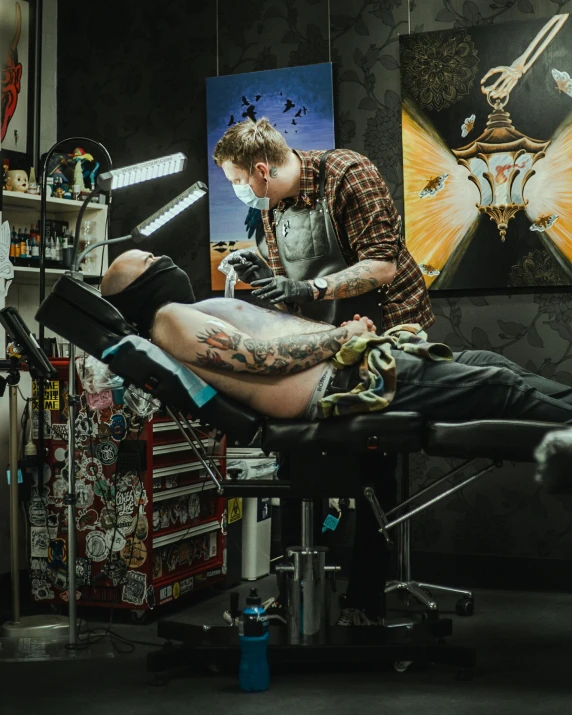 a tattooed man sits in a chair in his barbershop and watches the procedure