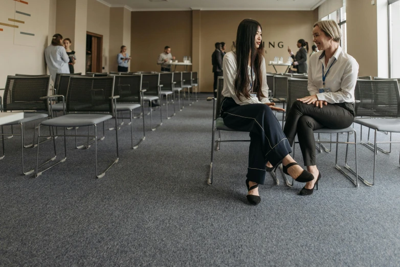 two girls are sitting on chairs talking in an open conference room