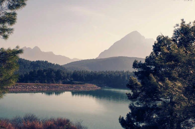 a tree lined landscape shows a small body of water and mountains behind it