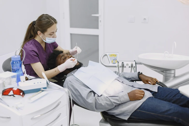 two women wearing face masks sit in the dentists chair while another lady in the dental room is holding a clipboard