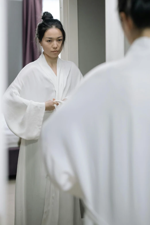 a woman stands in a room in front of her mirror, wearing a robe with a long flowing sleeve