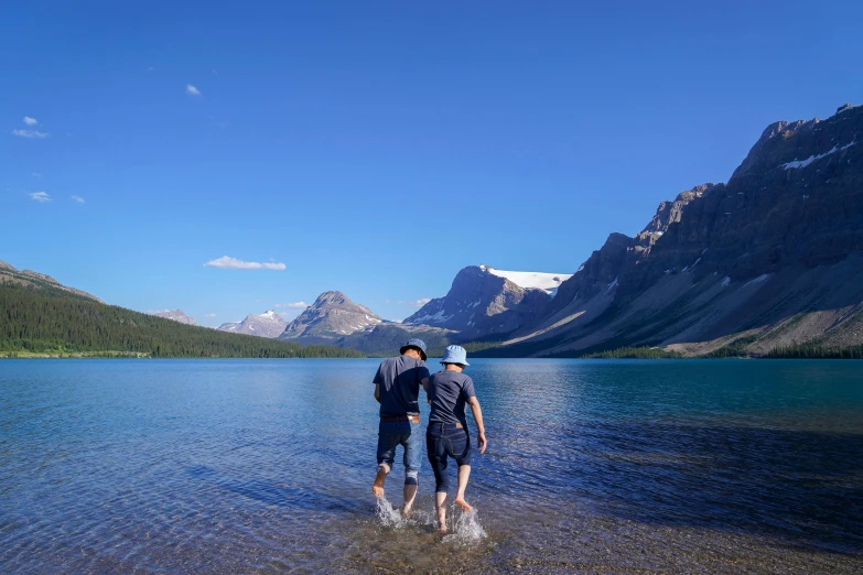 two men walking along side a river in front of mountains