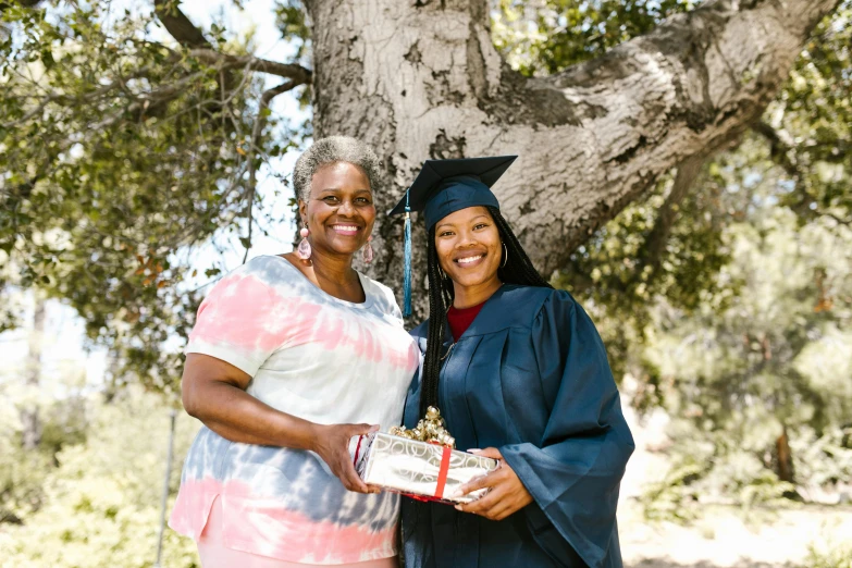 two woman in graduation attire pose under a large tree