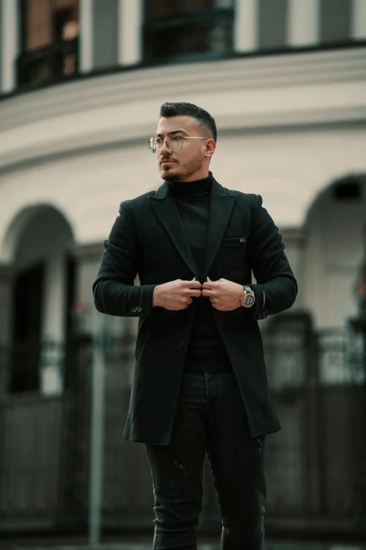 a man wearing glasses and a black jacket, standing in front of an old building