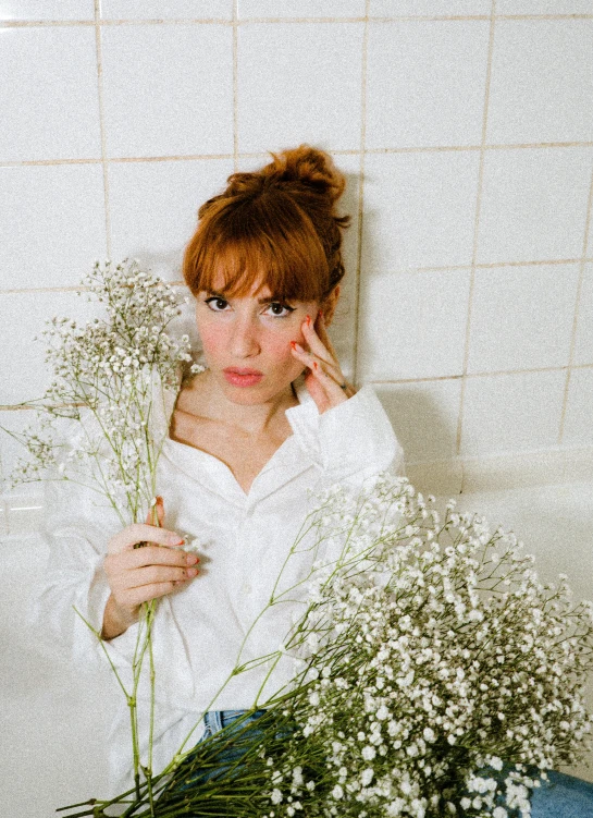 a woman wearing a white sweater laying on a white bed next to flowers