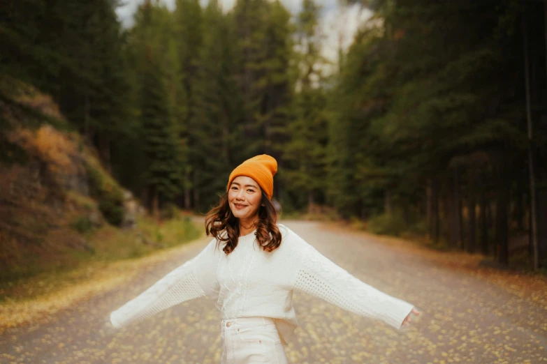 woman standing in front of a forest on a road wearing a orange beanie