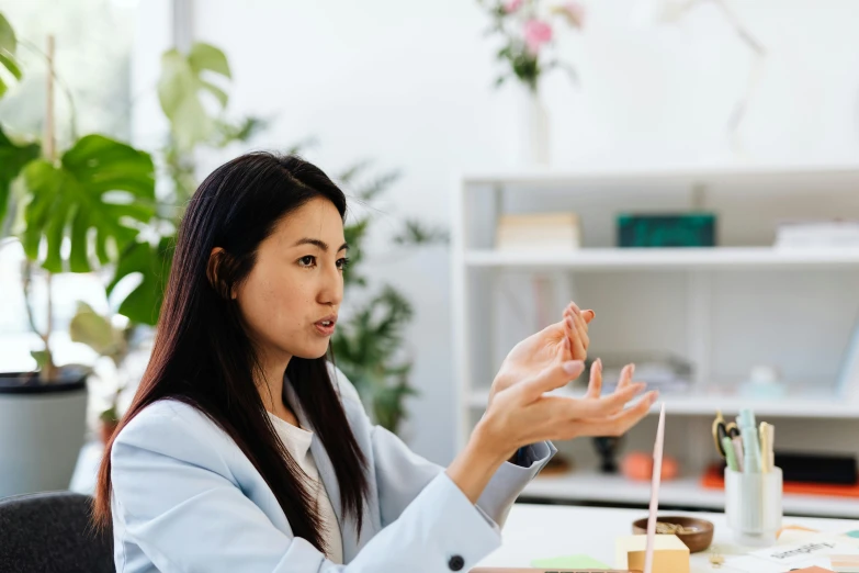 an asian woman wearing a jacket holds her hands together while sitting at a desk