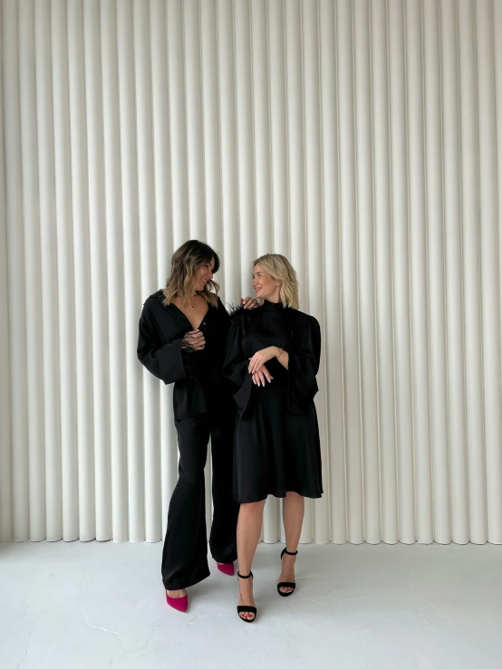 two women stand near a wall with vertical blinds