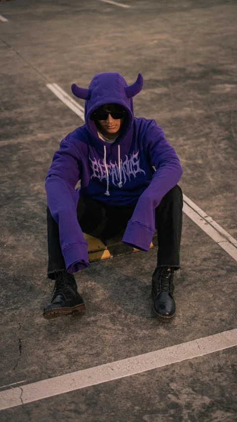 a young man sitting on the ground in a purple hoodie and mask