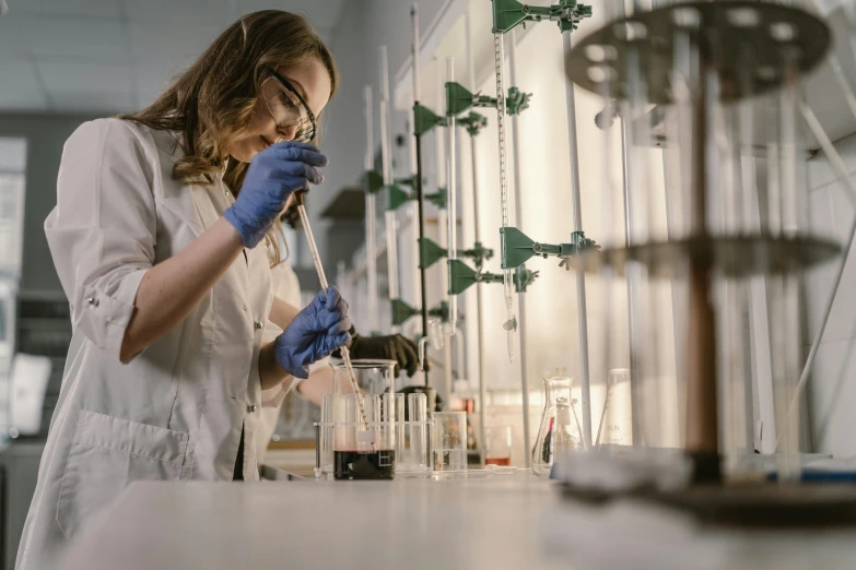 a young woman in a white lab coat and blue gloves holds a beakling for liquid