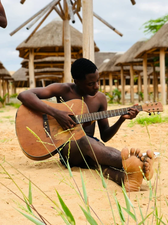 man sitting on sand playing guitar in front of huts