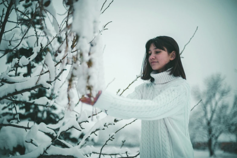 a girl in white is standing next to a tree in the snow