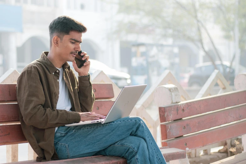 man on a laptop on a bench using his cell phone