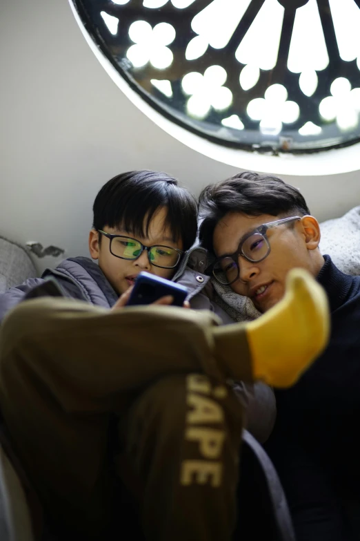 two boys are sitting on a couch one has glasses and the other is looking at a phone