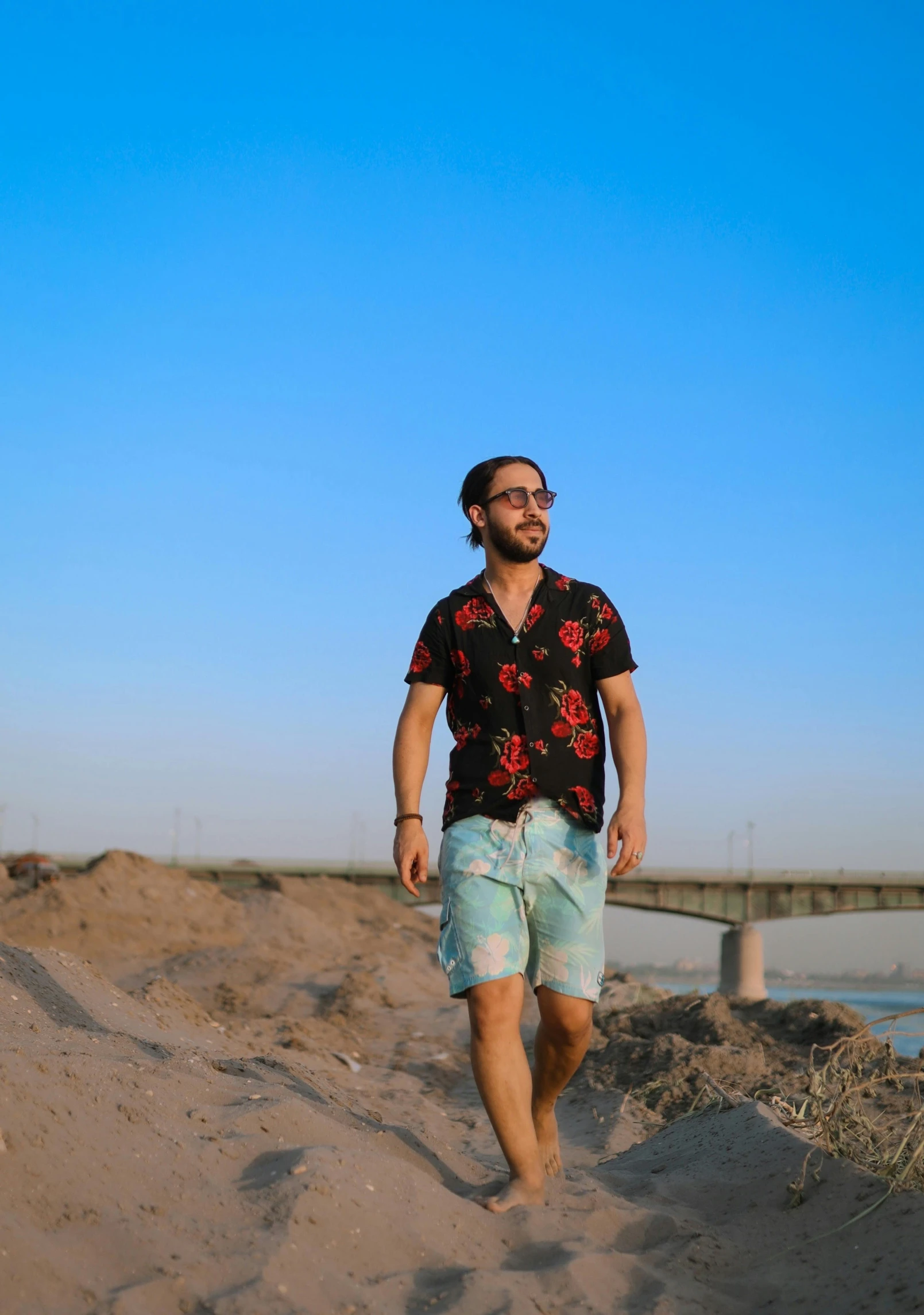 a man wearing shorts, a black shirt and sunglasses walks on the sand at the beach